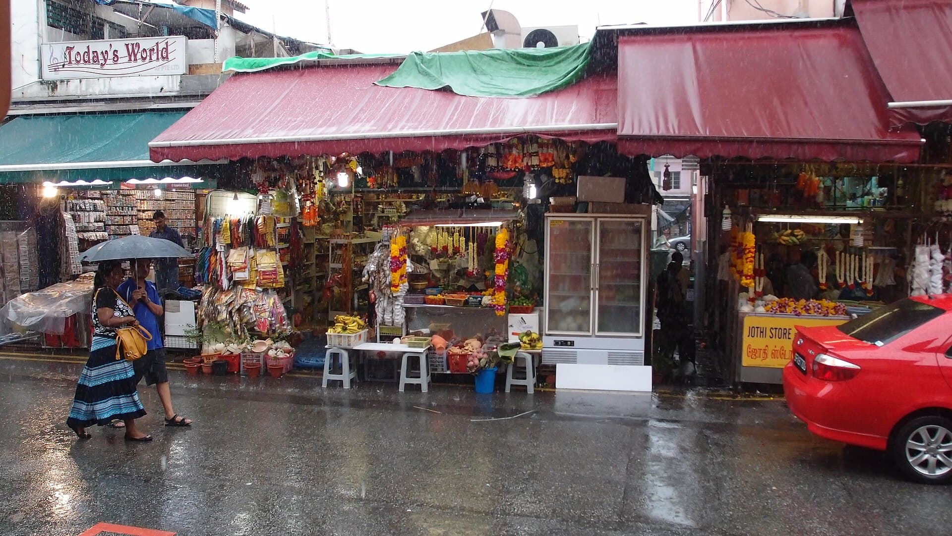 A wet street in Little India at night, with headlights and rain on the road.
